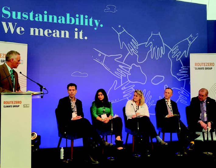 1 man stands at a podium with a microphone as 5 people sit to his left. The background reads 'Sustainability. We mean it.'