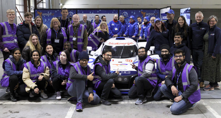 A group of people stand around a racecar. Many of the people are wearing purple high-vis jackets.