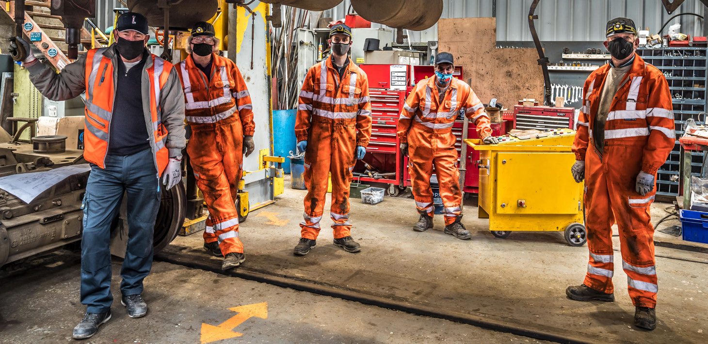 5 men in the team stand in the workshop, 4 are in orange high-vs jumpsuits and one is wearing an orange high-vis vest.