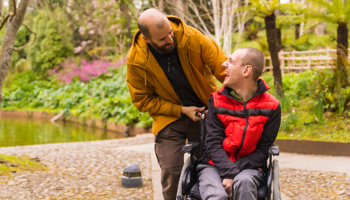 Two people laughing outdoors. One is using a wheelchair and the other is helping them.