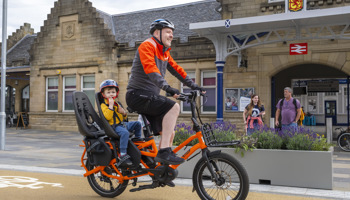 A man cycling with a boy in a child passenger seat, on a cycle path outside a mainline railway station